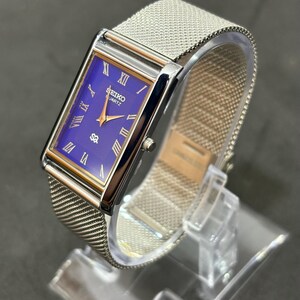 May include: A rectangular Seiko Quartz watch with a mesh metal band. The watch face is a deep blue with gold-colored hands and Roman numeral hour markers. The watch is displayed on a clear plastic stand.