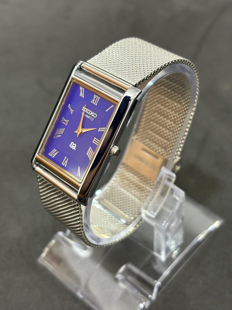 May include: A rectangular Seiko Quartz watch with a mesh metal band. The watch face is a deep blue with gold-colored hands and Roman numeral hour markers. The watch is displayed on a clear plastic stand.
