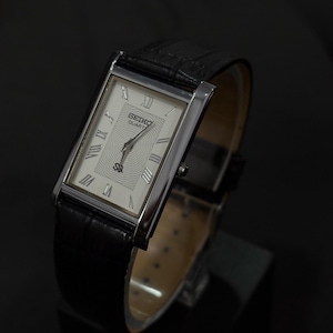 May include: A rectangular Seiko Quartz watch with a silver case and a black leather band. The watch face is white with Roman numerals and a textured center. The watch is displayed on a black surface.