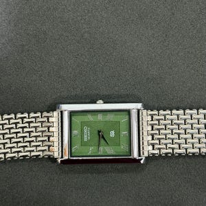 May include: A rectangular Seiko Quartz watch with a green face and silver-tone band. The watch face features Roman numerals and the Seiko logo. The band has a woven design.