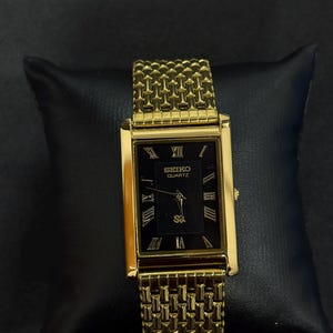 May include: A gold-toned Seiko Quartz watch with a rectangular black face and Roman numeral hour markers. The watch features a gold mesh band and is displayed on a black surface.