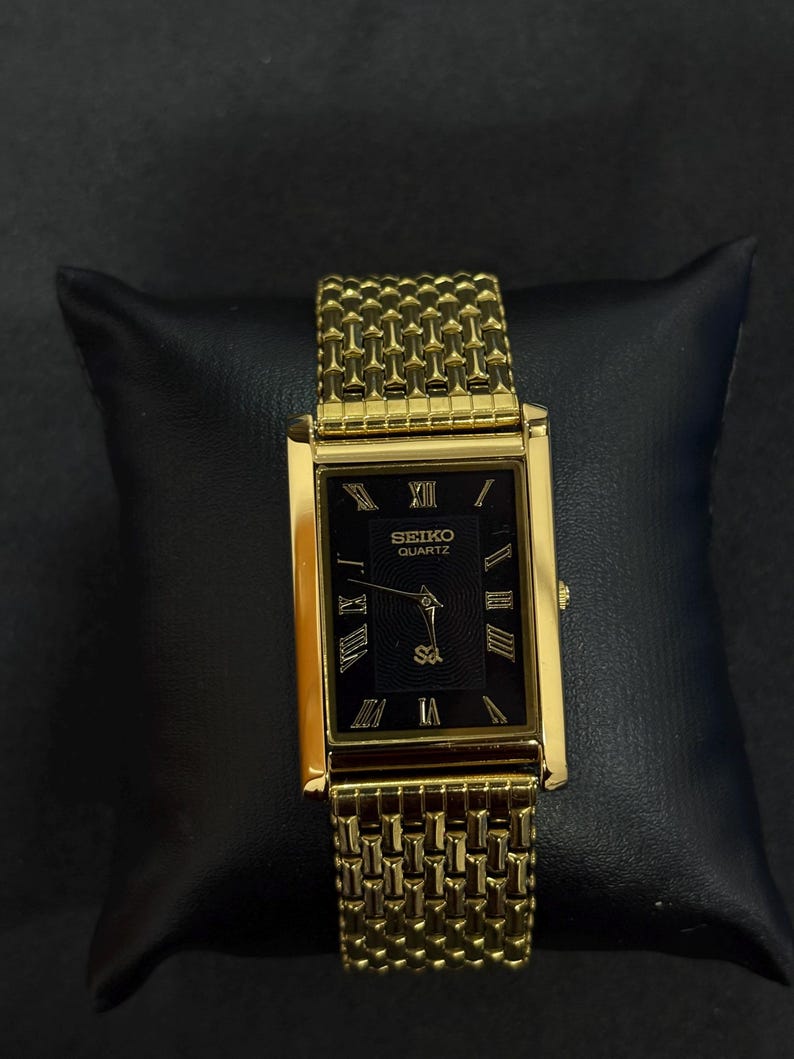 May include: A gold-toned Seiko Quartz watch with a rectangular black face and Roman numeral hour markers. The watch features a gold mesh band and is displayed on a black surface.
