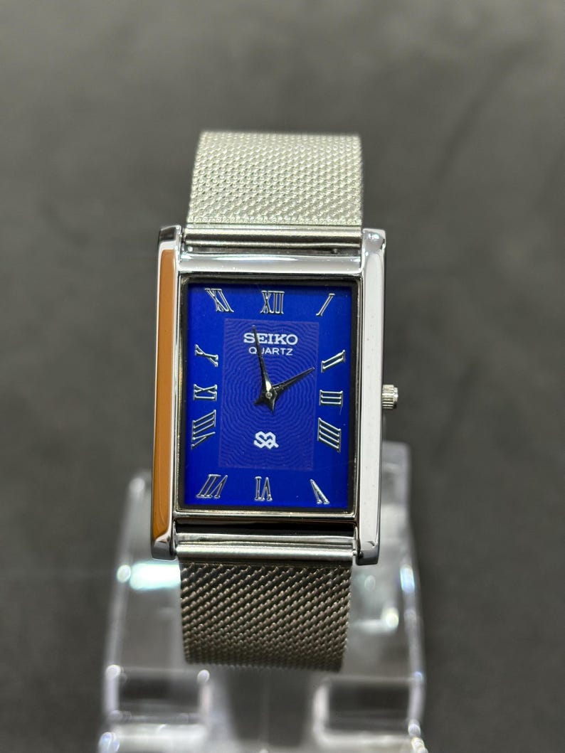 May include: A rectangular Seiko Quartz watch with a silver mesh band. The watch face is a vibrant blue with silver Roman numerals and hands. The watch case is silver, and the watch is displayed on a clear stand.