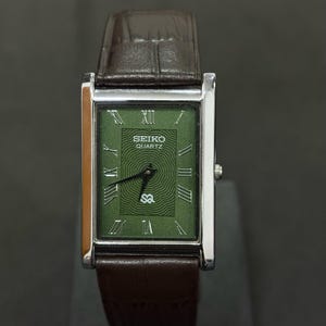 Może przedstawiać: Prostokątny zegarek Seiko Quartz z zieloną tarczą i srebrną kopertą. Zegarek posiada rzymskie cyfry i brązowy sk&oacute;rzany pasek. Na tarczy zegarka widnieje napis "SEIKO QUARTZ" oraz litery "SQ".