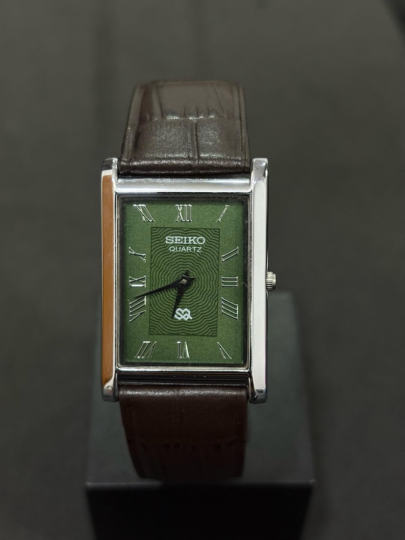 Może przedstawiać: Prostokątny zegarek Seiko Quartz z zieloną tarczą i srebrną kopertą. Zegarek posiada rzymskie cyfry i brązowy sk&oacute;rzany pasek. Na tarczy zegarka widnieje napis "SEIKO QUARTZ" oraz litery "SQ".