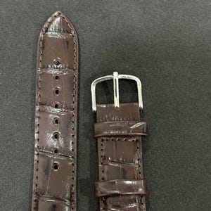 May include: Close-up of two sections of a brown watch strap. The strap is made of a textured material, possibly leather, with a crocodile pattern. The buckle is silver-coloured. The strap has several holes for adjustment.