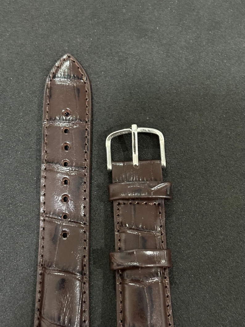 May include: Close-up of two sections of a brown watch strap. The strap is made of a textured material, possibly leather, with a crocodile pattern. The buckle is silver-coloured. The strap has several holes for adjustment.