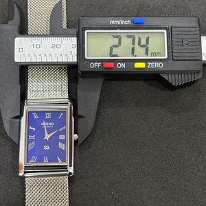 May include: A rectangular Seiko Quartz watch with a blue face and silver mesh band is measured with a digital caliper. The watch face displays Roman numerals and the Seiko logo. The measurement reads 27.4 mm.
