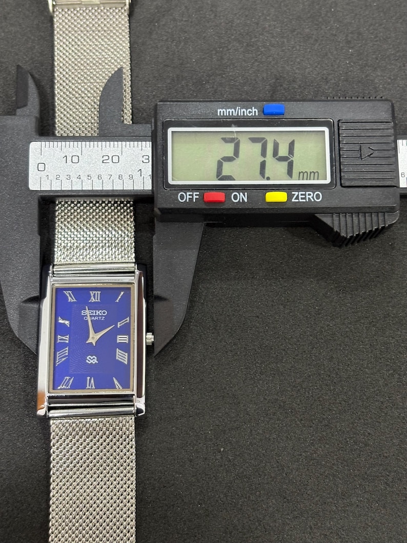 May include: A rectangular Seiko Quartz watch with a blue face and silver mesh band is measured with a digital caliper. The watch face displays Roman numerals and the Seiko logo. The measurement reads 27.4 mm.