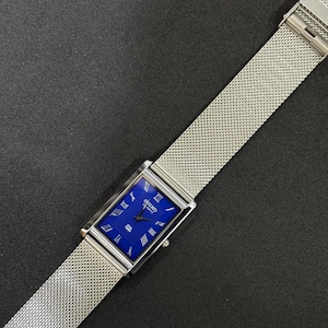 May include: A rectangular Seiko Quartz watch with a vibrant blue face and silver Roman numeral hour markers. The watch features a silver mesh band and a silver case. The watch is displayed on a dark surface.