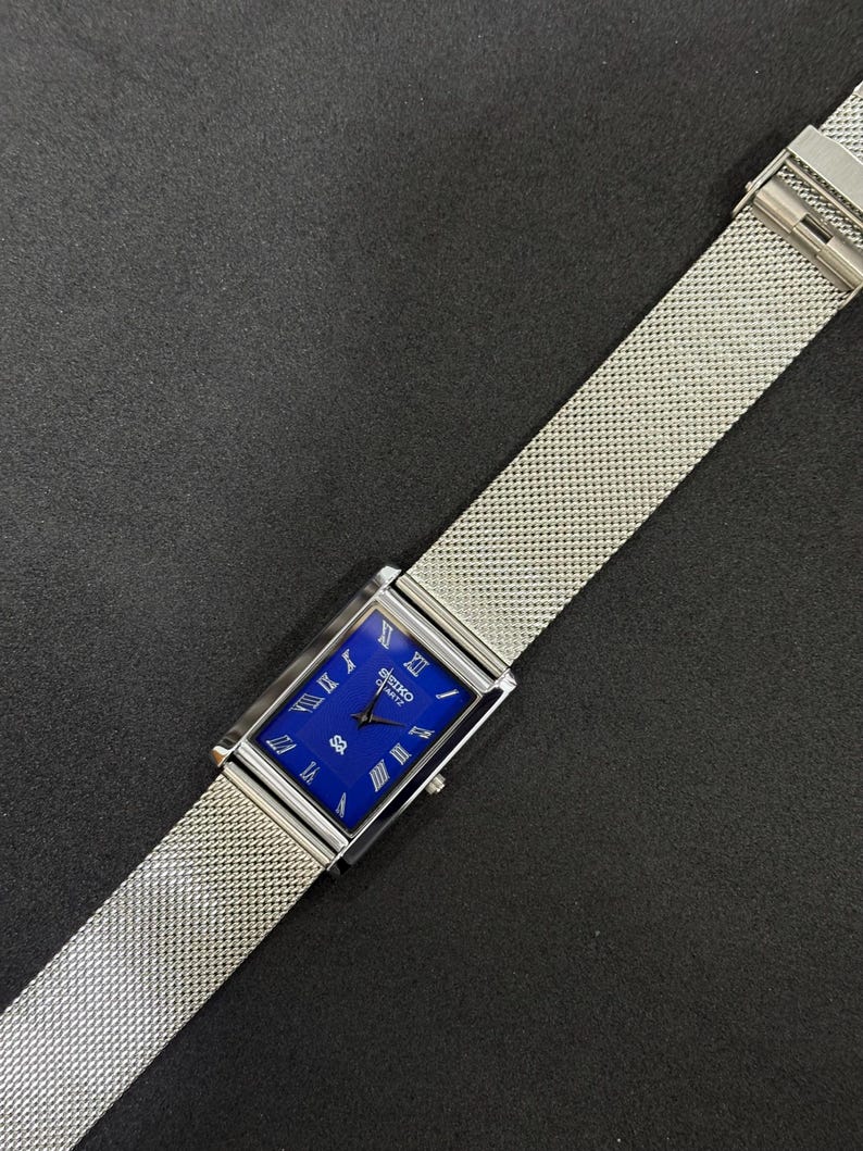 May include: A rectangular Seiko Quartz watch with a vibrant blue face and silver Roman numeral hour markers. The watch features a silver mesh band and a silver case. The watch is displayed on a dark surface.