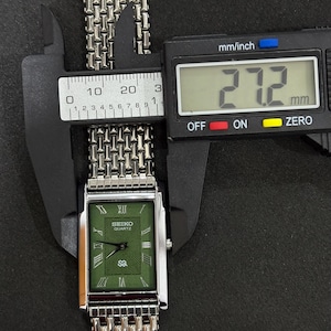 May include: A silver Seiko Quartz watch with a green rectangular face and a silver mesh band. The watch face displays Roman numerals and the brand name. The watch's width is measured at 27.2 mm using a digital caliper.