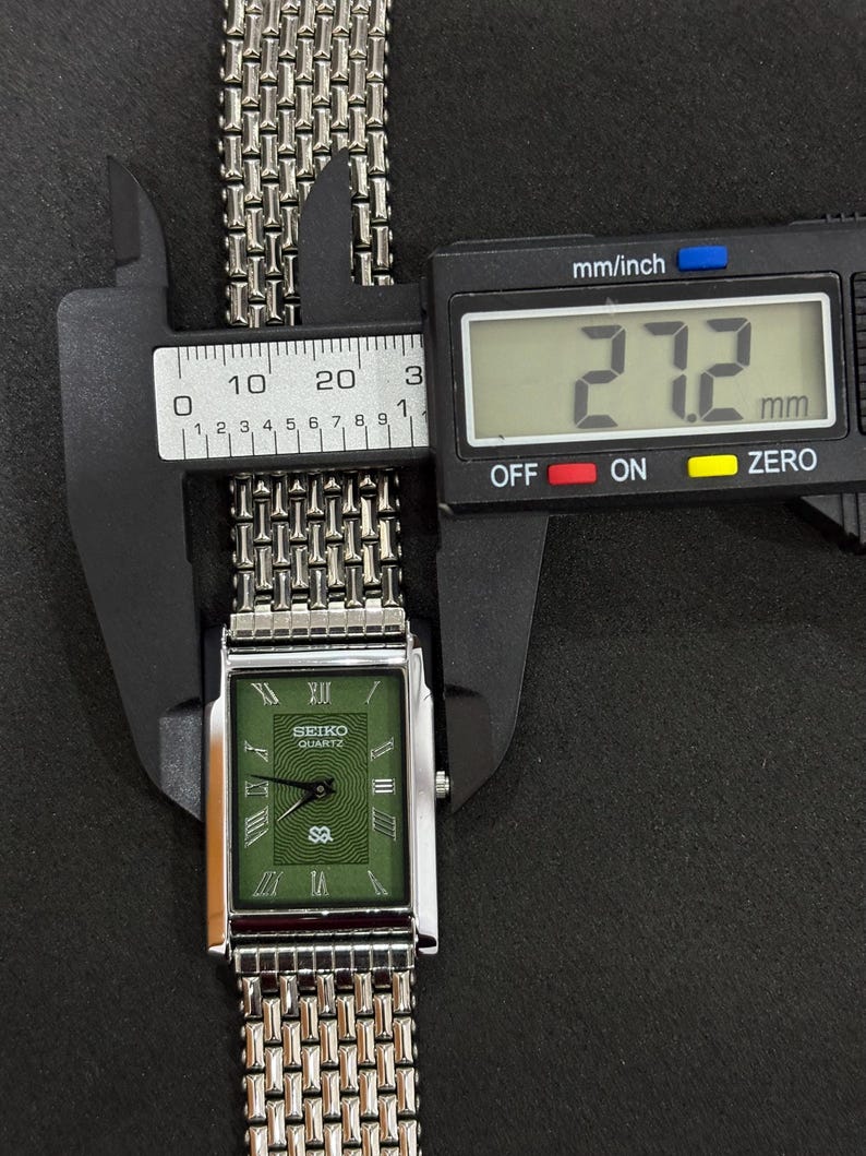 May include: A silver Seiko Quartz watch with a green rectangular face and a silver mesh band. The watch face displays Roman numerals and the brand name. The watch's width is measured at 27.2 mm using a digital caliper.