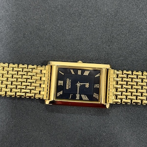 May include: A gold-toned Seiko Quartz watch with a rectangular black face and gold Roman numeral hour markers. The watch has a gold link band. The watch face displays the Seiko logo and the word "Quartz".