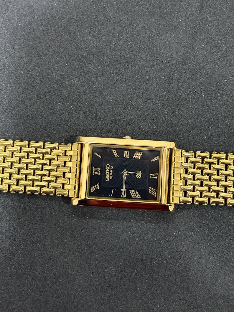 May include: A gold-toned Seiko Quartz watch with a rectangular black face and gold Roman numeral hour markers. The watch has a gold link band. The watch face displays the Seiko logo and the word "Quartz".