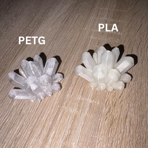 May include: Two crystal cluster sculptures, one silver and one white, sit on a wooden surface. The silver cluster is labeled "PETG" and the white cluster is labeled "PLA".
