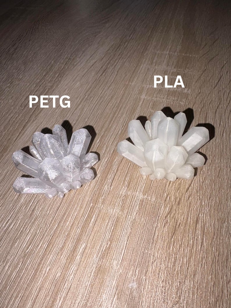 May include: Two crystal cluster sculptures, one silver and one white, sit on a wooden surface. The silver cluster is labeled "PETG" and the white cluster is labeled "PLA".