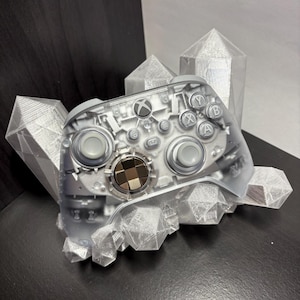 May include: A clear Xbox controller with visible internal components, resting on a silver, geometric crystal-like structure. The controller has gray buttons and a metallic center. The background features additional silver crystal formations.