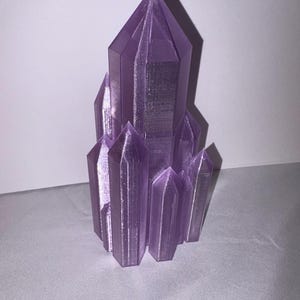 K&ouml;nnte beinhalten: Ein Cluster aus lila Kristallformationen, mit einem zentralen, gro&szlig;en Kristall, der von kleineren umgeben ist. Die Kristalle haben ein facettiertes Design, das das Licht einf&auml;ngt und Schatten erzeugt. Das Objekt steht vor einem schlichten Hintergrund.