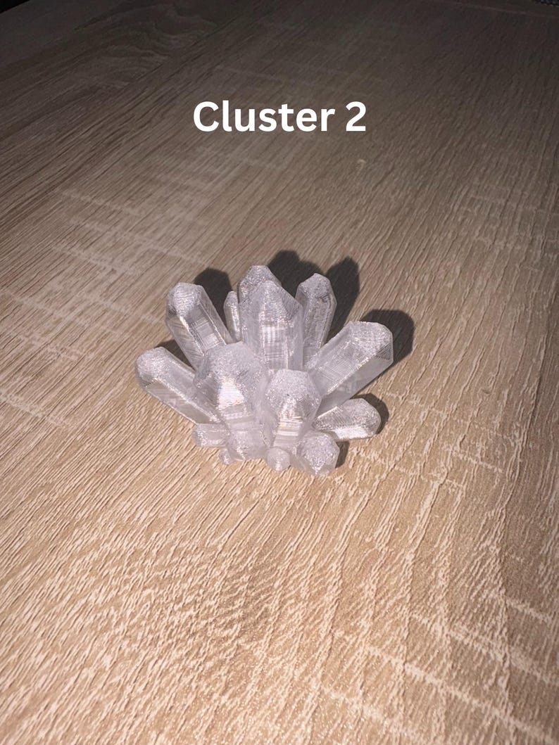 May include: A clear crystal cluster with multiple points, resting on a wooden surface. The text "Cluster 2" is displayed in white letters above the crystal. The crystal is a light, translucent color.