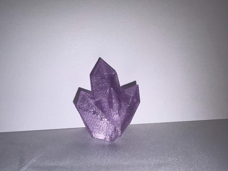 K&ouml;nnte beinhalten: Ein Cluster aus lila Kristallformationen. Die Kristalle haben scharfe, spitze Kanten und eine strukturierte Oberfl&auml;che. Die Farbe ist ein leuchtendes, schimmerndes Lila, und die Gesamtform ist symmetrisch und ansprechend.