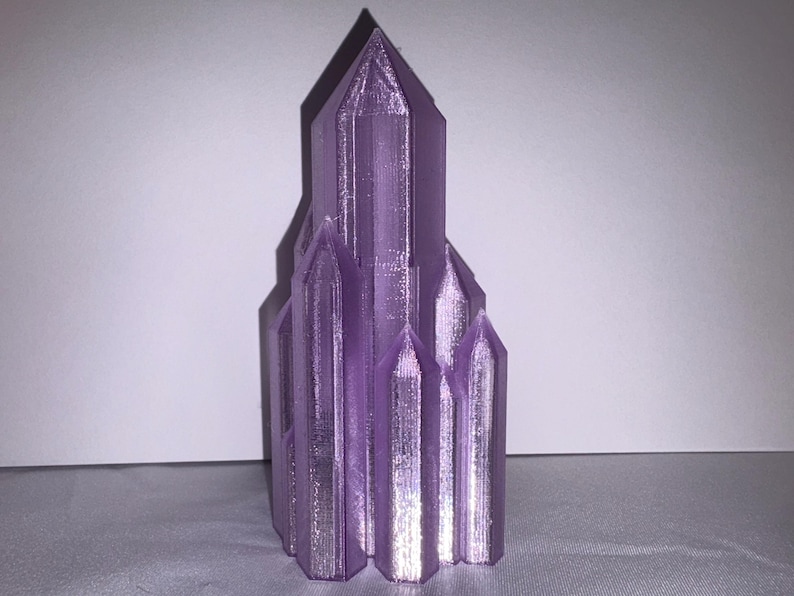 K&ouml;nnte beinhalten: Eine 3D-gedruckte, lila Kristallskulptur. Die Skulptur hat ein kathedralenartiges Design mit mehreren T&uuml;rmen und einer strukturierten Oberfl&auml;che. Die Farbe ist ein leuchtendes, metallisches Lila, und das Objekt steht vor einem neutralen Hintergrund.