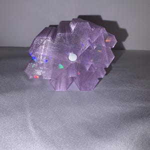 K&ouml;nnte beinhalten: Eine lavendelfarbene, geometrische Skulptur mit strukturierter Oberfl&auml;che. Die Skulptur hat einen zentralen wei&szlig;en Kreis und weist schillernde Glanzlichter auf. Das Objekt befindet sich vor einem neutralen Hintergrund.