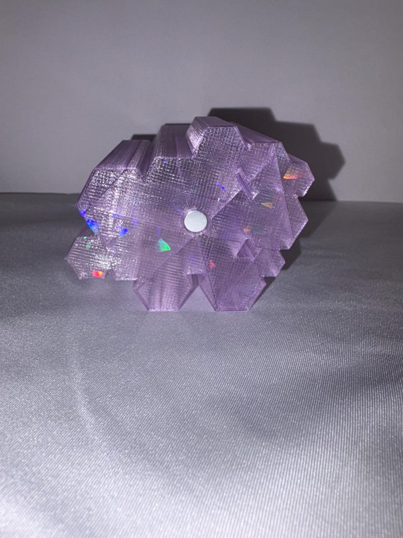 K&ouml;nnte beinhalten: Eine lavendelfarbene, geometrische Skulptur mit strukturierter Oberfl&auml;che. Die Skulptur hat einen zentralen wei&szlig;en Kreis und weist schillernde Glanzlichter auf. Das Objekt befindet sich vor einem neutralen Hintergrund.