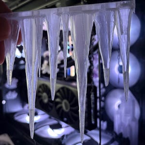May include: A clear, 3D-printed object resembling a row of icicles. The icicles are suspended from a horizontal bar and taper to sharp points. The background shows computer components with illuminated fans, creating a cool, wintery theme.