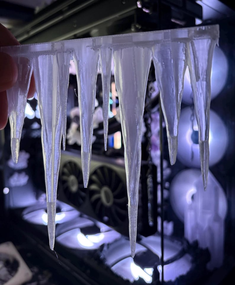 May include: A clear, 3D-printed object resembling a row of icicles. The icicles are suspended from a horizontal bar and taper to sharp points. The background shows computer components with illuminated fans, creating a cool, wintery theme.