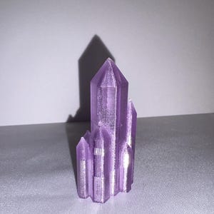 K&ouml;nnte beinhalten: Eine Ansammlung von lila Kristallformationen, jede mit einem spitzen, facettierten Design. Die Kristalle sind in einer Gruppe angeordnet, mit einem gr&ouml;&szlig;eren zentralen Kristall und mehreren kleineren. Die Kristalle sind etwa 10 cm hoch.