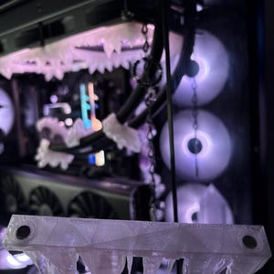 May include: A clear, 3D-printed decorative piece resembling icicles, held in front of a computer case with glowing fans. The piece has a geometric design with pointed icicle shapes hanging from the bottom. The computer case has a black frame.