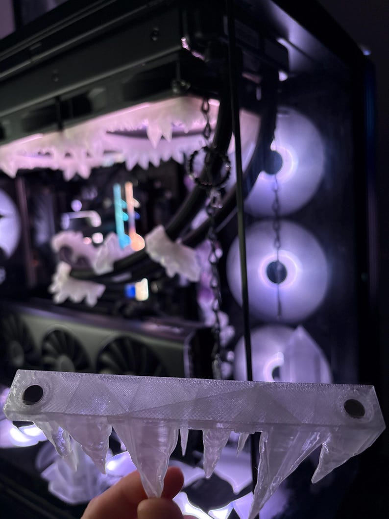 May include: A clear, 3D-printed decorative piece resembling icicles, held in front of a computer case with glowing fans. The piece has a geometric design with pointed icicle shapes hanging from the bottom. The computer case has a black frame.