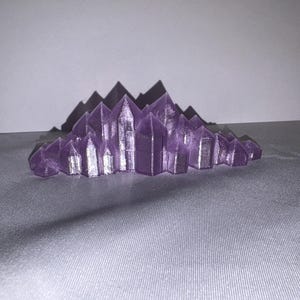 K&ouml;nnte beinhalten: Eine lila Kristallcluster-Skulptur mit scharfen, spitzen Formationen. Die Kristalle sind in einer geschichteten, berg&auml;hnlichen Struktur angeordnet. Die Skulptur besteht aus einem festen Material und steht auf einer grauen Oberfl&auml;che.