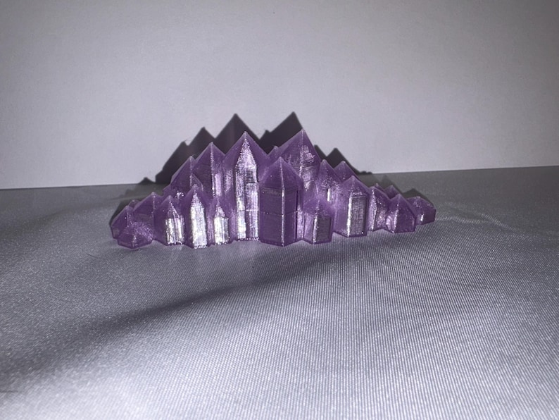 K&ouml;nnte beinhalten: Eine lila Kristallcluster-Skulptur mit scharfen, spitzen Formationen. Die Kristalle sind in einer geschichteten, berg&auml;hnlichen Struktur angeordnet. Die Skulptur besteht aus einem festen Material und steht auf einer grauen Oberfl&auml;che.