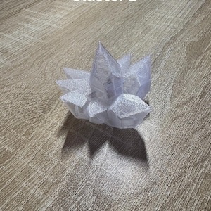 May include: A clear, crystalline cluster with a pointed top and multiple facets. The cluster is white and sits on a wooden surface. The text "Cluster 1" is visible in the upper left corner.