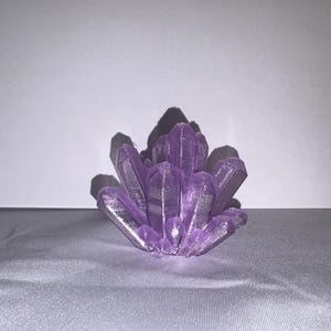 K&ouml;nnte beinhalten: Ein Cluster aus violetten Kristallspitzen, das einer Geode oder Quarzformation &auml;hnelt. Die Kristalle sind in einem strahlenf&ouml;rmigen Muster angeordnet, mit einer strukturierten Oberfl&auml;che und einem metallischen Glanz. Das Objekt ist vor einem neutralen Hintergrund platziert.