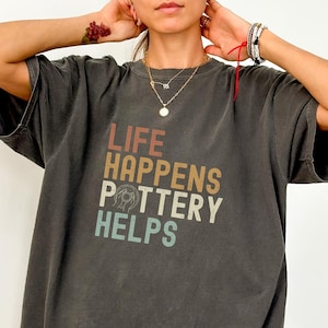 May include: Dark gray t-shirt with the text "LIFE HAPPENS POTTERY HELPS" in a retro font. The shirt has a relaxed fit and short sleeves. The shirt is a dark gray color.