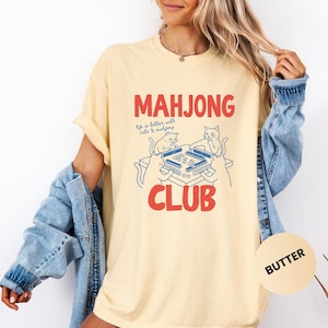 K&ouml;nnte beinhalten: Hellgelbes T-Shirt mit roter und blauer Grafik. Die Grafik zeigt "MAHJONG CLUB" mit dem Satz "life is better with cats & mahjong" und zeigt zwei Katzen beim Mahjong-Spielen. Das T-Shirt hat kurze &Auml;rmel und eine lockere Passform.