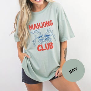 K&ouml;nnte beinhalten: Hellgr&uuml;nes T-Shirt mit den roten Worten "MAHJONG CLUB" und einer blauen Illustration von Katzen, die Mahjong spielen. Das Shirt hat auch den Text "life is better with cats & mahjong".