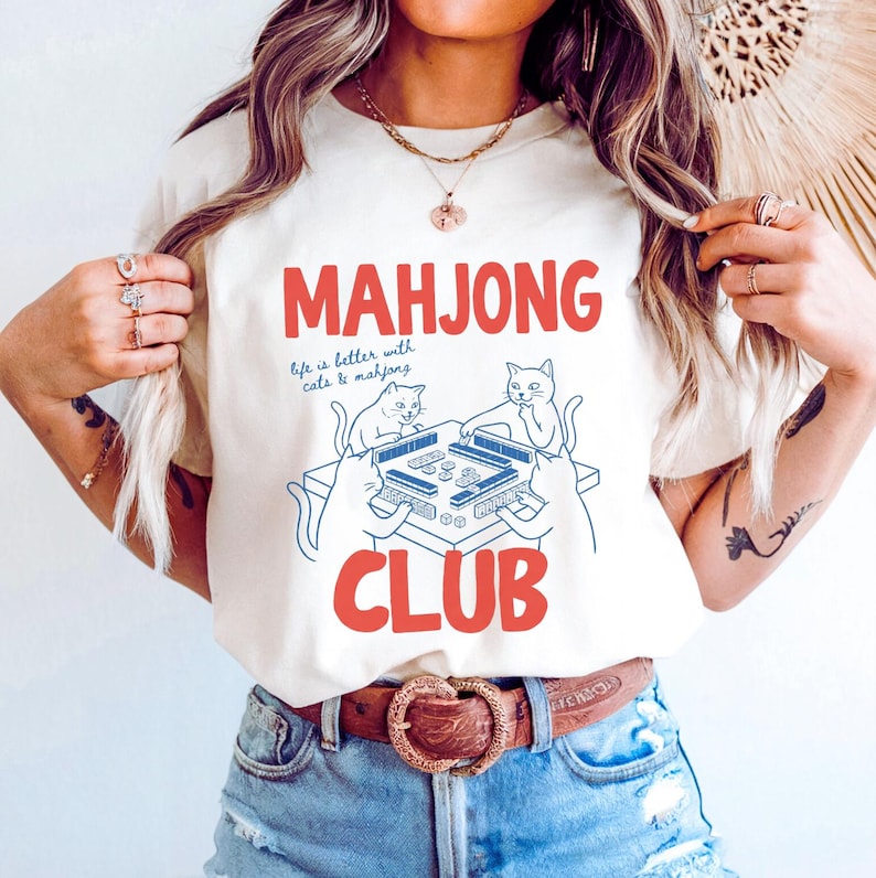 K&ouml;nnte beinhalten: Cremefarbenes T-Shirt mit den roten Worten "MAHJONG CLUB" und dem Satz "life is better with cats & mahjong" darunter. Das Design zeigt zwei Katzen beim Mahjong-Spielen. Das Shirt wird mit einer blauen Jeans und einem braunen G&uuml;rtel getragen.