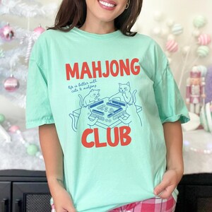 K&ouml;nnte beinhalten: Mintgr&uuml;nes T-Shirt mit den Worten "MAHJONG CLUB" in roten Blockbuchstaben. Unter dem Wort "MAHJONG" steht der Satz "life is better with cats & mahjong". Eine blaue Illustration von Katzen, die Mahjong spielen, ist abgebildet.