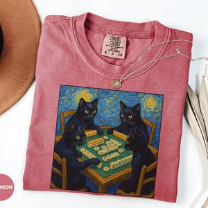 May include: Crimson Comfort Colors t-shirt featuring a painting of four black cats playing mahjong, set against a starry night backdrop. The shirt has a crew neck and short sleeves. A silver necklace with a small pendant is draped on the shirt.