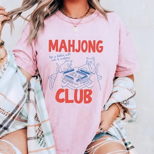 K&ouml;nnte beinhalten: Hellrosa T-Shirt mit dem roten Schriftzug "MAHJONG CLUB". Unter dem Wort "MAHJONG" steht der Satz "life is better with cats & mahjong". Eine blaue Illustration von Katzen, die Mahjong spielen, befindet sich unter dem Text.