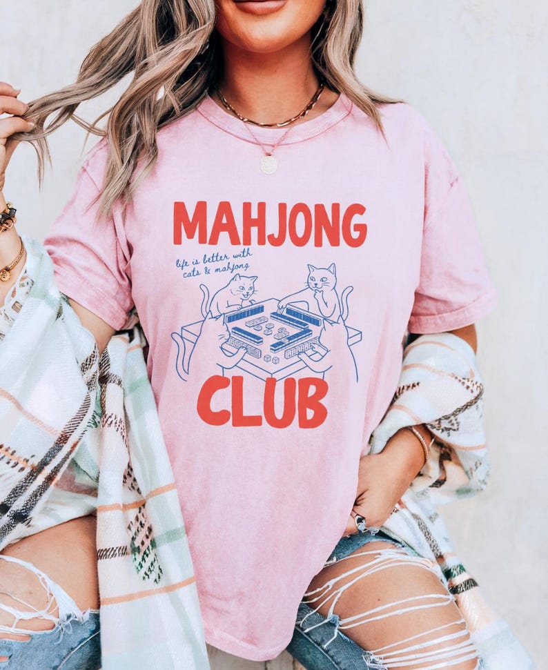 K&ouml;nnte beinhalten: Hellrosa T-Shirt mit dem roten Schriftzug "MAHJONG CLUB". Unter dem Wort "MAHJONG" steht der Satz "life is better with cats & mahjong". Eine blaue Illustration von Katzen, die Mahjong spielen, befindet sich unter dem Text.