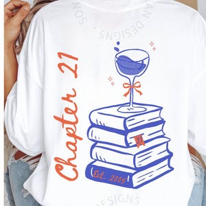 May include: White t-shirt with a graphic design featuring a wine glass atop a stack of blue books. The text "Chapter 21" is in orange, and "Est. 2005" is printed on the books. The design includes a red ribbon and sparkles.