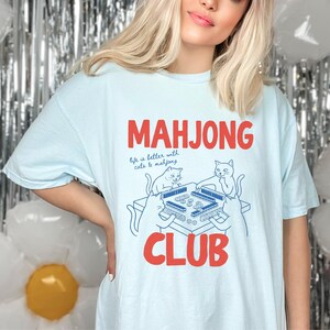 K&ouml;nnte beinhalten: Hellblaues T-Shirt mit den Worten "MAHJONG CLUB" in roten Blockbuchstaben. Das Shirt zeigt eine Grafik eines Mahjong-Tisches mit zwei Katzen und dem Text "life is better with cats & mahjong."