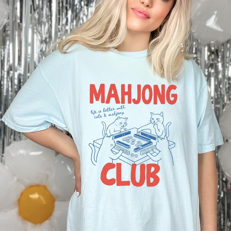 K&ouml;nnte beinhalten: Hellblaues T-Shirt mit den Worten "MAHJONG CLUB" in roten Blockbuchstaben. Das Shirt zeigt eine Grafik eines Mahjong-Tisches mit zwei Katzen und dem Text "life is better with cats & mahjong."