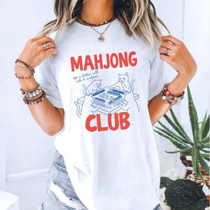 K&ouml;nnte beinhalten: Wei&szlig;es T-Shirt mit den roten Worten "MAHJONG CLUB" und einer blauen Illustration von Katzen, die Mahjong spielen. Der Text "life is better with cats & mahjong" ist ebenfalls auf dem Shirt.