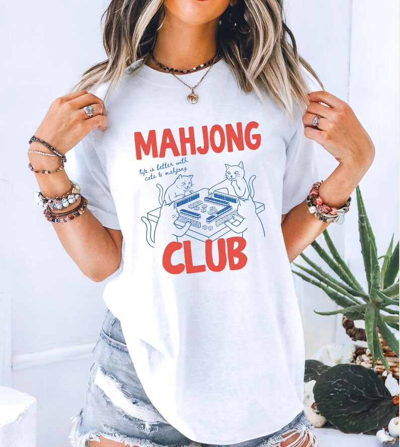 K&ouml;nnte beinhalten: Wei&szlig;es T-Shirt mit den roten Worten "MAHJONG CLUB" und einer blauen Illustration von Katzen, die Mahjong spielen. Der Text "life is better with cats & mahjong" ist ebenfalls auf dem Shirt.